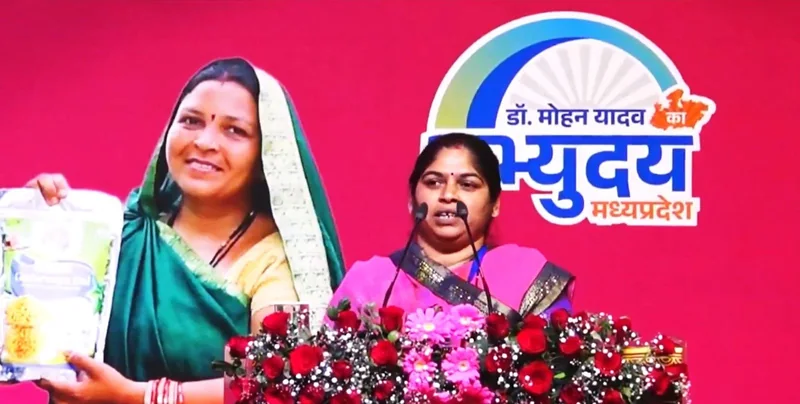 लाल पृष्ठभूमि वाले मंच पर माइक के सामने बोलती एक महिला; पीछे पैकेट पकड़े महिला की बड़ी तस्वीर